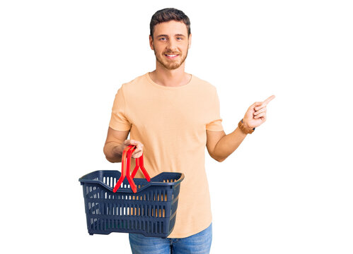 Handsome young man with bear holding supermarket shopping basket smiling happy pointing with hand and finger to the side