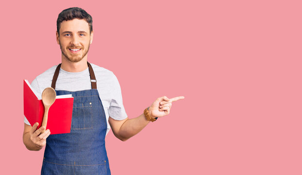 Handsome young man with bear wearing professional baker apron reading cooking recipe book smiling happy pointing with hand and finger to the side