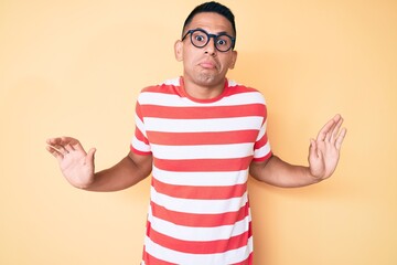 Young handsome latin man wearing casual clothes and glasses clueless and confused expression with...