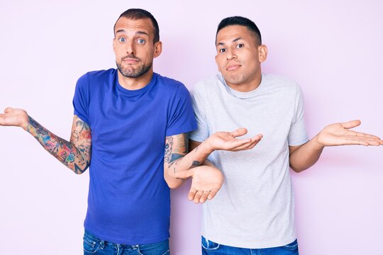 Young gay couple of two men wearing casual clothes clueless and confused expression with arms and hands raised. doubt concept.