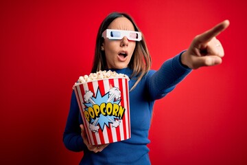 Young blonde woman wearing 3d glasses and eating pack of popcorn watching a movie on cinema Pointing with finger surprised ahead, open mouth amazed expression, something on the front