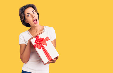 Young beautiful woman holding gift scared and amazed with open mouth for surprise, disbelief face