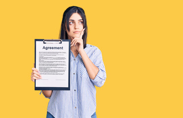 Young beautiful girl holding clipboard with agreement document serious face thinking about question...