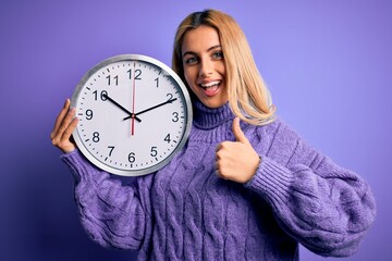 Young beautiful blonde woman doing countdown holding big clock over purple background happy with big smile doing ok sign, thumb up with fingers, excellent sign