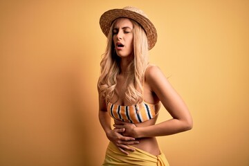 Young beautiful blonde woman on vacation wearing bikini and hat over yellow background with hand on...