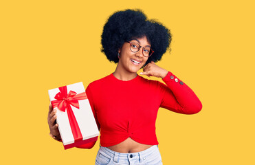 Young african american girl holding gift smiling doing phone gesture with hand and fingers like talking on the telephone. communicating concepts.