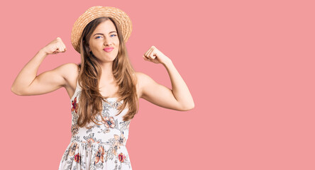 Beautiful caucasian young woman wearing summer hat showing arms muscles smiling proud. fitness concept.