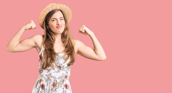 Beautiful caucasian young woman wearing summer hat showing arms muscles smiling proud. fitness concept.