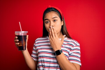 Young asian girl drinking cola fizzy refreshment using straw over isolated red background cover...