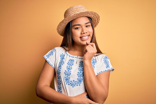 Young beautiful asian girl wearing casual t-shirt and hat standing over yellow background looking confident at the camera smiling with crossed arms and hand raised on chin. Thinking positive.