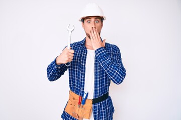Young handsome man weaing handyman uniform holding wrench covering mouth with hand, shocked and afraid for mistake. surprised expression