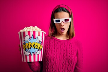Obraz premium Young beautiful girl watching movie using 3d glasses eating box with popcorns In shock face, looking skeptical and sarcastic, surprised with open mouth