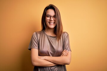Obraz premium Beautiful brunette girl wearing casual t-shirt and glasses over isolated yellow background happy face smiling with crossed arms looking at the camera. Positive person.