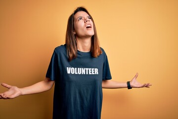 Young beautiful brunette girl doing volunteering wearing t-shirt with volunteer message word crazy and mad shouting and yelling with aggressive expression and arms raised. Frustration concept.
