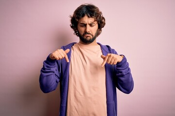 Young handsome sporty man with beard wearing casual sweatshirt over pink background Pointing down looking sad and upset, indicating direction with fingers, unhappy and depressed.