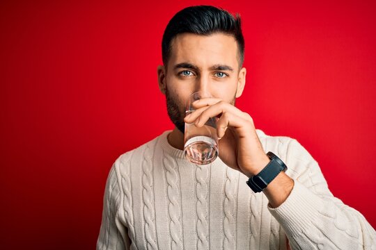 Young handsome man drinking glass of healthy water to refreshment standing over isolated red background