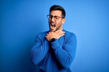 Young handsome man with beard wearing casual sweater and glasses over blue background shouting suffocate because painful strangle. Health problem. Asphyxiate and suicide concept.