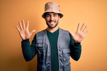 Handsome tourist man with beard on vacation wearing explorer hat over yellow background showing and pointing up with fingers number ten while smiling confident and happy.