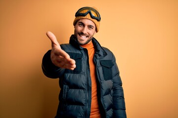 Young handsome skier man with beard wearing snow sportswear and ski goggles smiling friendly offering handshake as greeting and welcoming. Successful business.