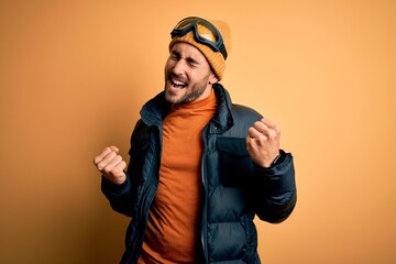 Young handsome skier man with beard wearing snow sportswear and ski goggles very happy and excited doing winner gesture with arms raised, smiling and screaming for success. Celebration concept.