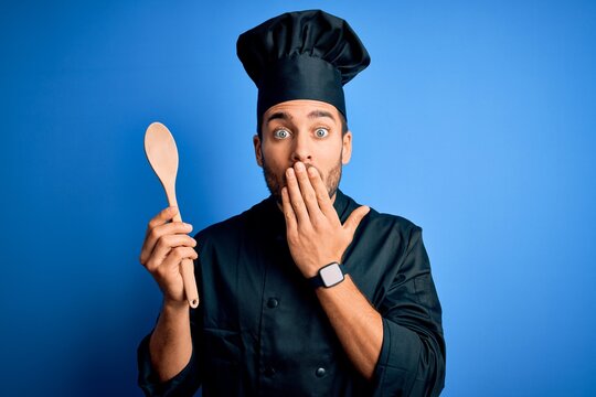 Young cooker man with beard wearing uniform holding wooden spoon over blue background cover mouth with hand shocked with shame for mistake, expression of fear, scared in silence, secret concept