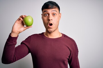 Young hispanic man eating fresh green apple as healthy nutrition over isolated background scared in shock with a surprise face, afraid and excited with fear expression