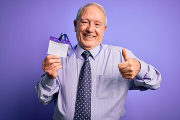 Senior grey haired business man holding identification tag over purple background happy with big smile doing ok sign, thumb up with fingers, excellent sign