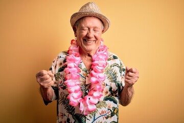 Grey haired senior man wearing summer hat and hawaiian lei over yellow background excited for...