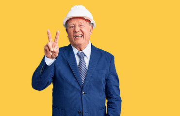 Senior handsome grey-haired man wearing suit and architect hardhat smiling with happy face winking at the camera doing victory sign. number two.