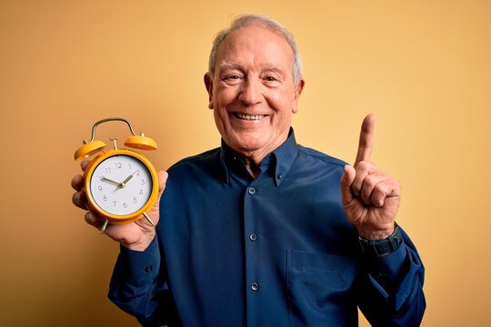 Senior grey haired man holding vintage alarm clock over yellow background surprised with an idea or question pointing finger with happy face, number one