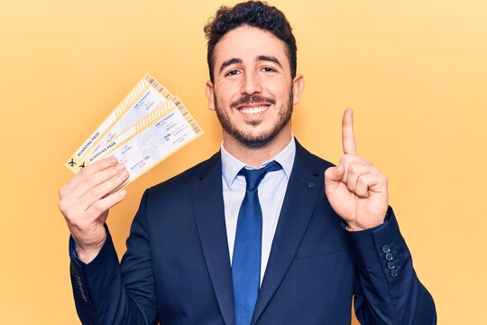 Young hispanic man wearing suit holding boarding pass smiling with an idea or question pointing finger with happy face, number one