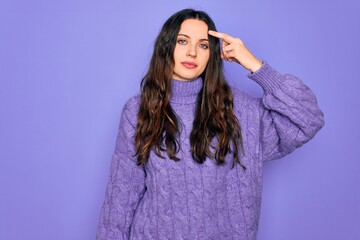 Young beautiful woman wearing casual turtleneck sweater standing over purple background pointing unhappy to pimple on forehead, ugly infection of blackhead. Acne and skin problem