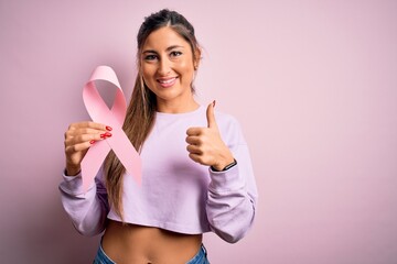 Young beautiful brunette woman holding pink cancer ribbon symbol over isolated background happy with big smile doing ok sign, thumb up with fingers, excellent sign