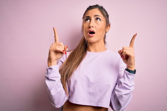 Young beautiful sport woman wearing sweatshirt over pink isolated background amazed and surprised looking up and pointing with fingers and raised arms.
