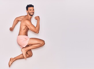Fototapeta premium Young handsome hispanic man on vacation wearing swimwear shirtless smiling happy. Jumping with smile on face celebrating with fists up over isolated white background