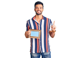 Young handsome hispanic man holding empty frame surprised with an idea or question pointing finger with happy face, number one