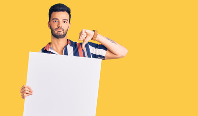 Young handsome hispanic man holding cardboard banner with blank space with angry face, negative sign showing dislike with thumbs down, rejection concept