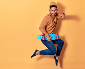 Young handsome latin man wearing wool cap jumping with smile on face. Holding skate over isolated yellow background