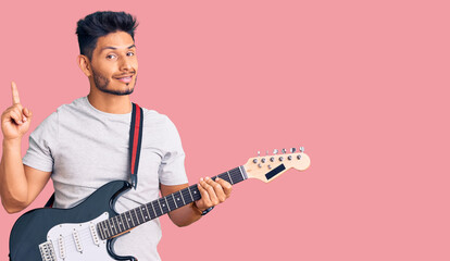 Handsome latin american young man playing electric guitar surprised with an idea or question pointing finger with happy face, number one
