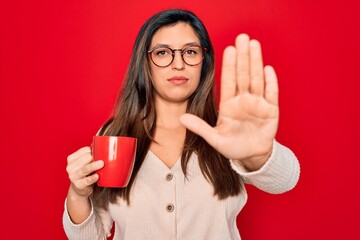 Young hispanic woman wearing glasses drinking a cup of coffee over red background with open hand doing stop sign with serious and confident expression, defense gesture