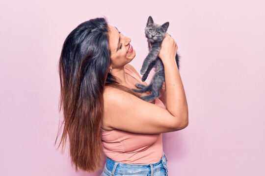 Young beautiful latin woman smilling happy. Standing with smile on face holding adorable cat over isolated pink background