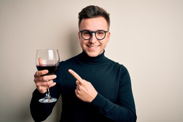 Young handsome caucasian man drinking an alcoholic glass of red wine over isolated background very happy pointing with hand and finger