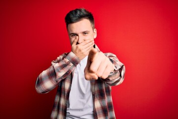 Young handsome caucasian man wearing casual modern shirt over red isolated background laughing at you, pointing finger to the camera with hand over mouth, shame expression