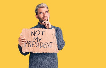 Young handsome blond man holding not my president cardboard banner serious face thinking about...