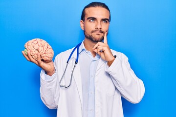 Young handsome man wearing doctor coat holding brain serious face thinking about question with hand...