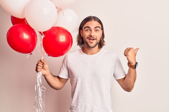 Young handsome man holding balloons pointing thumb up to the side smiling happy with open mouth