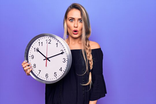 Young beautiful blonde woman doing countdown using big clock over purple background scared and amazed with open mouth for surprise, disbelief face