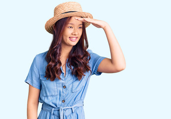 Young beautiful chinese girl wearing summer hat very happy and smiling looking far away with hand over head. searching concept.
