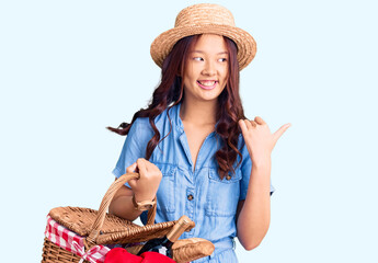Young beautiful chinese girl wearing summer hat and holding picnic wicker basket with bread pointing thumb up to the side smiling happy with open mouth