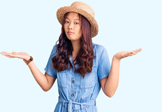Young beautiful chinese girl wearing summer hat clueless and confused expression with arms and hands raised. doubt concept.